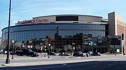 The wide, convex facade of a glassy sports arena and the parking lot, lawn, and lampposts in front of it. The words "Xcel Energy Center" sit in red atop the arena.