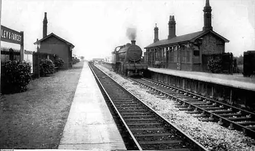A black and white photo of two railway tracks with a platform either side. On the right-hand track is a locomotive producing steam. Each platform has shrubberies and a platform building, and on the left-hand platform there is a cut-off sign that reads "LEY & FARCET".