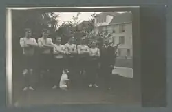 This black and white image is of James Arbuthnot and his 1911 Oregon Agricultural College wrestling team, and his dog. There are six shirtless men, all with arms crossed, lined up in front of a bush, with a three-story building in the background. Coach Arbuthnot, dressed in a suit, stands at the far right. His dog sits on its hind legs, front paws in the air in the begging position facing towards the coach. The dog is white save for a dark patch around its right eye.