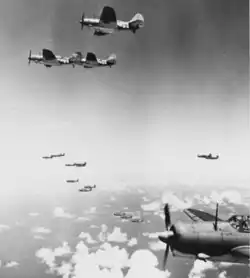 16 planes in profile flying to the right. Cumulus clouds and ocean at the bottom of the picture.
